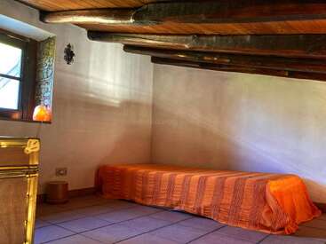This cozy attic room features a low wooden ceiling with exposed beams, a single bed covered in an orange blanket, a window, and minimal rustic decor.