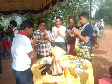 A group of people stands around a table covered with yellow cloth, hands in prayer position, participating in a traditional outdoor ceremony, surrounded by trees and observers.