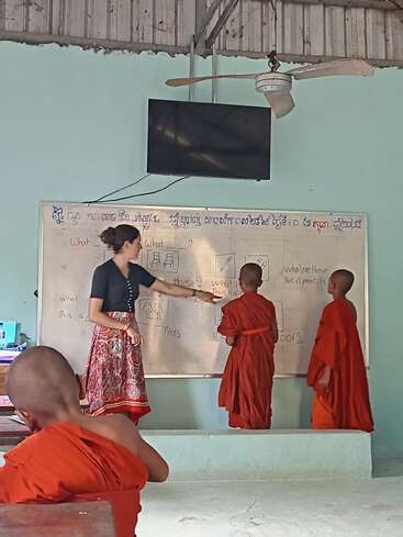 A teacher in a patterned skirt instructs young monks in orange robes at a whiteboard, engaging in an English lesson inside a classroom with simple decor.