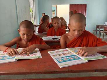 Young Buddhist monks, dressed in orange robes, sit at wooden desks in a classroom, intently reading colorful textbooks, deeply engaged in their studies together.