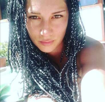 Eine Frau mit langen, schwarz-weiß geflochtenen Haaren und einem Nasenring schaut im Freien in die Kamera, während das Sonnenlicht ihr Gesicht und den Hintergrund beleuchtet.