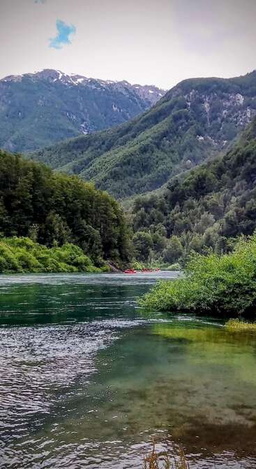 Ein ruhiger Fluss fließt durch üppige grüne Wälder mit hoch aufragenden Bergen im Hintergrund. Rote Flöße treiben sanft, umgeben von ruhiger natürlicher Schönheit und klarem Wasser.