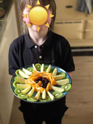 A child, face covered with a sun emoji, holds a colorful fruit plate with apple slices, kiwi rounds, orange segments, and cherries arranged in a circular pattern.