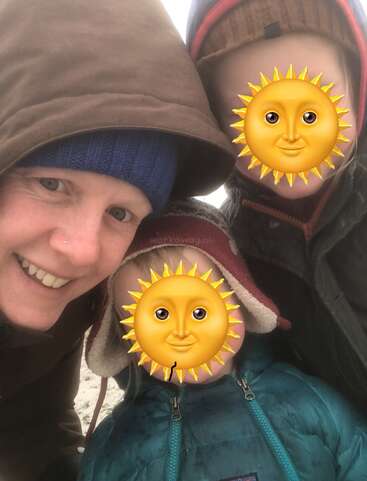 Three people pose closely outdoors, dressed warmly in winter jackets and hats. Two faces are covered with sun emojis, while one adult smiles directly at the camera.
