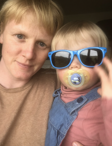 A smiling adult holds a young child with light hair. The child wears blue sunglasses and a pacifier, dressed in overalls and a pink shirt.