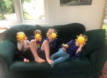 Four children are sitting comfortably on a dark green couch near large windows. Their faces are covered with sun emoticons. The atmosphere appears casual and relaxed.