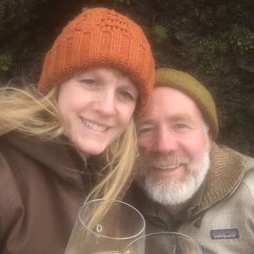 Una pareja sonriente, con gorros de abrigo, posa muy cerca el uno del otro al aire libre, con unas bebidas en la mano. La mujer tiene el pelo rubio y el hombre lleva barba. Un momento acogedor y alegre que comparten juntos.