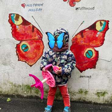 A child stands in front of a mural with butterfly wings, appearing to have wings and a butterfly mask. The wall is decorated with colorful butterfly art.