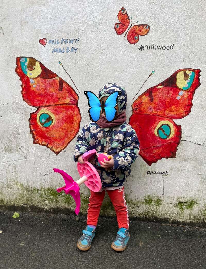 A child stands against a mural of butterfly wings, perfectly aligned to appear as their own wings. The child holds a pink toy and wears colorful clothes.