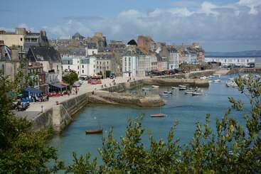 The image depicts a picturesque coastal town with a row of buildings and a body of water, featuring a prominent boat or ship in the foreground, surrounded by a natural landscape.