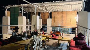 A cozy rooftop lounge at night, featuring cushioned seating, low tables, bamboo blinds, warm lighting, and two people relaxing and using their phones in a laid-back atmosphere.