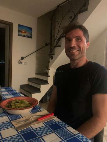Un hombre sonriente está sentado a una mesa con motivos azules con un plato de pasta, un tenedor y una servilleta. Una escalera de caracol y obras de arte decoran la acogedora habitación.