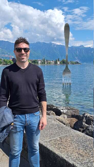 Um homem sorridente, de óculos escuros, está à beira de um lago, com montanhas ao fundo. Uma escultura gigante em forma de garfo emerge da água atrás dele, sob um céu azul.