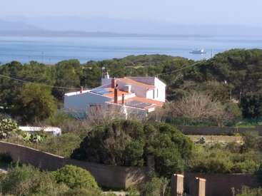 Uma casa branca com telhado de terracota fica em meio a árvores verdes, com vista para um mar calmo onde se avista um navio ao longe. A cena transmite uma sensação de paz e serenidade.