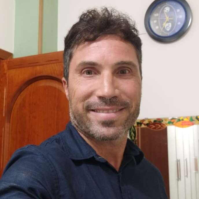 Um cara com cabelo curto e escuro e barba, vestindo uma camisa azul-marinho, sorri para uma selfie em uma sala com móveis de madeira e um relógio de parede.