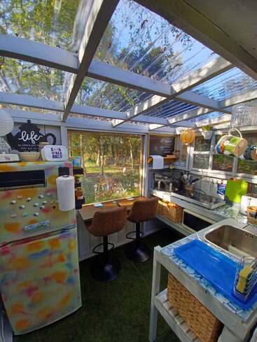 Une cuisine chaleureuse et baignée de soleil, avec un toit en verre, un réfrigérateur coloré, des sièges au comptoir près d'une fenêtre, une décoration vivante et un cadre extérieur verdoyant, créant une atmosphère lumineuse et accueillante.