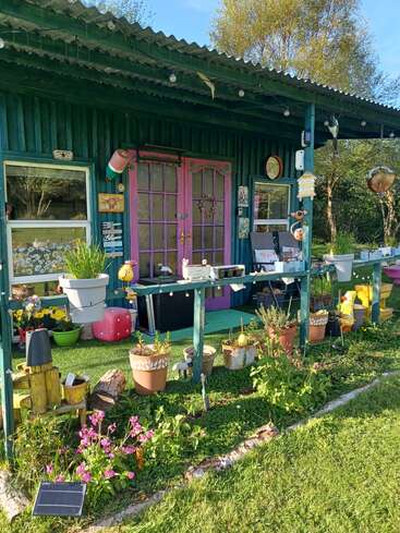 Une charmante cabane de jardin aux murs bleu sarcelle et aux portes roses, entourée de plantes en pot, de décorations de jardin, de fleurs et d'éléments de décoration rustiques, dégage une atmosphère fantaisiste et créative.