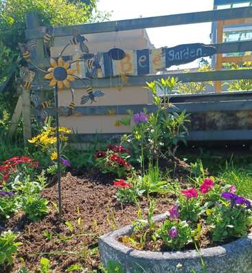 A vibrant bee-themed garden with colorful flowers, a decorative sunflower and bee spinner, lush greenery, and a sign reading "Bee Garden" attached to a fence.