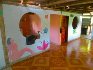 Une pièce colorée avec un sol carrelé, des fenêtres circulaires, une porte rouge et des murs peints représentant des fleurs et une femme nue assise à côté d'une grande fleur rose.