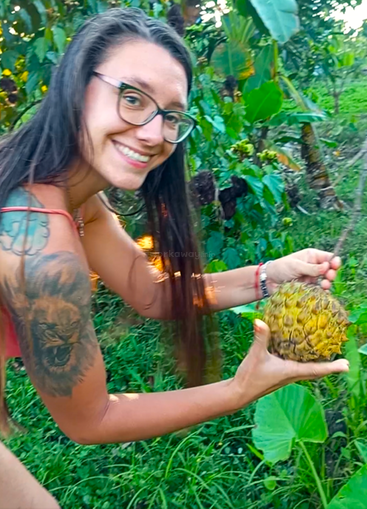 Une femme souriante, portant des lunettes et arborant un tatouage de lion, récolte un fruit dans un jardin luxuriant et verdoyant, entouré de plantes tropicales et de la lumière du soleil.