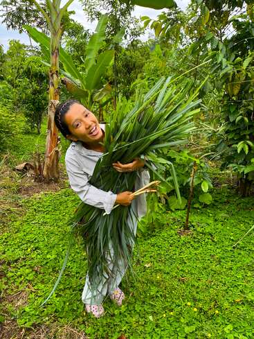 Une femme joyeuse se tient dans un jardin verdoyant, tenant un gros paquet de longues feuilles fraîches. Elle sourit largement, entourée d'un feuillage vibrant.