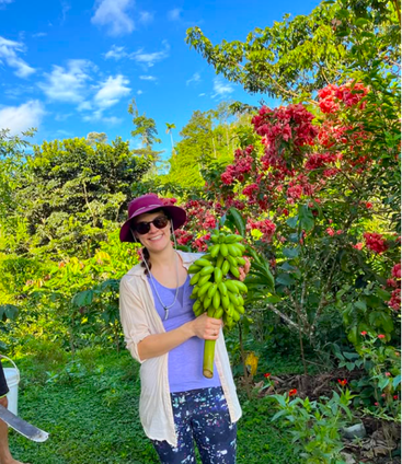 Une femme souriante portant des lunettes de soleil et un chapeau se tient dans un jardin vibrant et luxuriant, tenant un gros régime de bananes vertes, entouré de verdure et de fleurs colorées.
