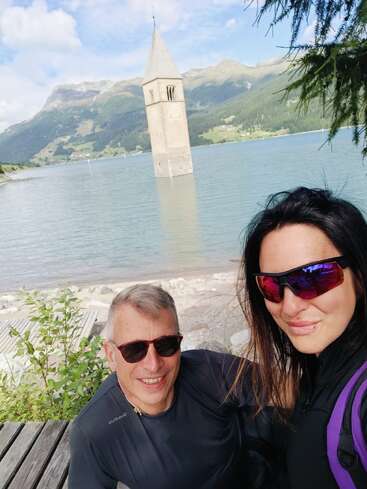 Un hombre y una mujer con gafas de sol sonríen para hacerse un selfi junto a un lago en el que se ve sumergido un campanario, rodeados de montañas y vegetación bajo un cielo azul.