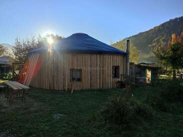 Une construction ronde en bois, de style yourte, se dresse dans une zone herbeuse, baignée par la lumière du soleil qui filtre à travers les arbres, entourée de montagnes et de nature, évoquant la paix et la simplicité.