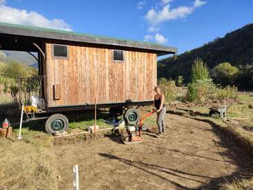 Une femme utilise un compacteur de sol à côté d'une petite maison en bois sur roues, entourée par la campagne, les arbres, les montagnes et un ciel bleu limpide.