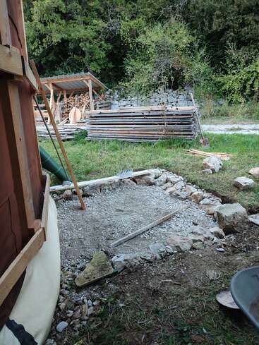 Un chantier de construction avec une fondation en gravier, entouré de pierres et d'outils. À l'arrière-plan, on aperçoit des piles de bois et un petit hangar. Une végétation luxuriante.