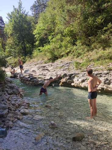 Trois personnes profitent d'un ruisseau limpide et rocailleux dans une forêt luxuriante. L'une d'elles plonge dans l'eau, une autre observe la scène, tandis que la troisième se détend sur les rochers à proximité.