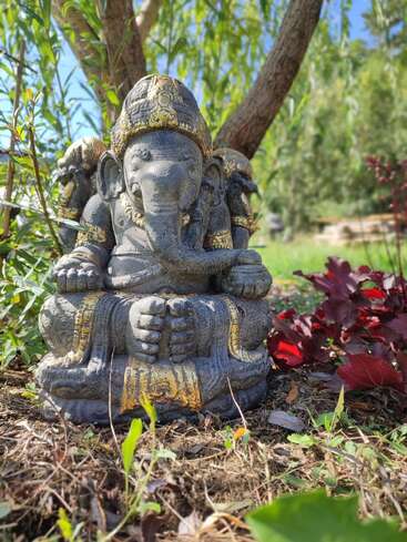 Une statue en pierre représentant Ganesha trône gracieusement dans un jardin luxuriant, entourée de verdure et de plantes rouges, ornée de touches dorées, sous un arbre ombragé baigné de soleil.