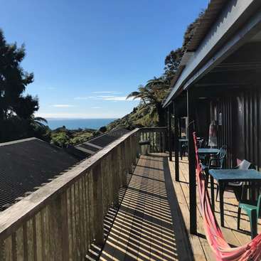 The image depicts a serene wooden deck with a railing, overlooking the ocean, featuring tables and chairs, surrounded by lush trees and a clear blue sky on a sunny day.