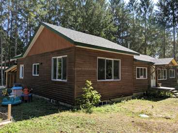 A cozy wooden cabin sits in a sunny clearing surrounded by tall pine trees. Large windows and a small porch add charm to this rustic retreat.