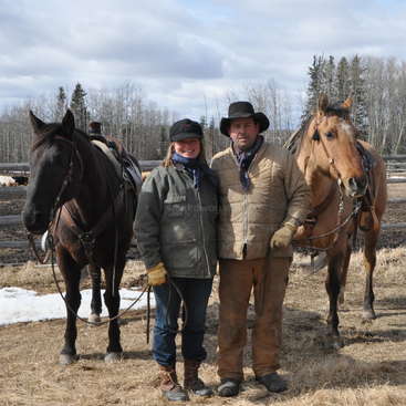 Das Bild zeigt einen Mann und eine Frau, die vor zwei Pferden stehen. Der Mann trägt einen Cowboyhut und die Frau ist winterlich gekleidet.