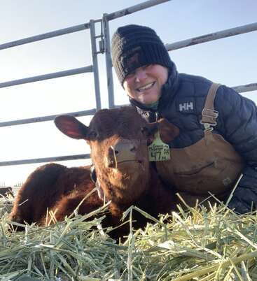 A smiling person dressed warmly sits beside a brown calf lying on hay. They are inside a fenced pen. The calf has an ear tag numbered 47.