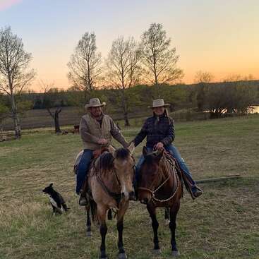 Dos personas con sombreros de vaquero montan a caballo uno al lado del otro al atardecer en un campo abierto, acompañados por un perro blanco y negro cercano. Un campo tranquilo.