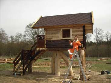 A person in an orange jumpsuit paints a wooden treehouse elevated on large logs, with a ladder nearby, surrounded by trees and open grassland.