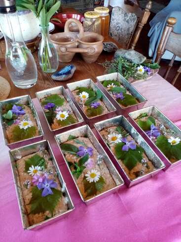Eight rectangular baking tins filled with dough, beautifully decorated with green leaves and colorful edible flowers, sit on a pink tablecloth. Rustic kitchen items surround them.