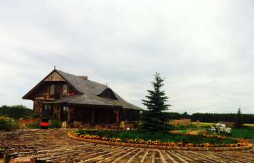 A rustic wooden house sits in a rural landscape with a circular stone pathway, colorful flowerbeds, green lawn, pine tree, and white outdoor chairs under a cloudy sky.
