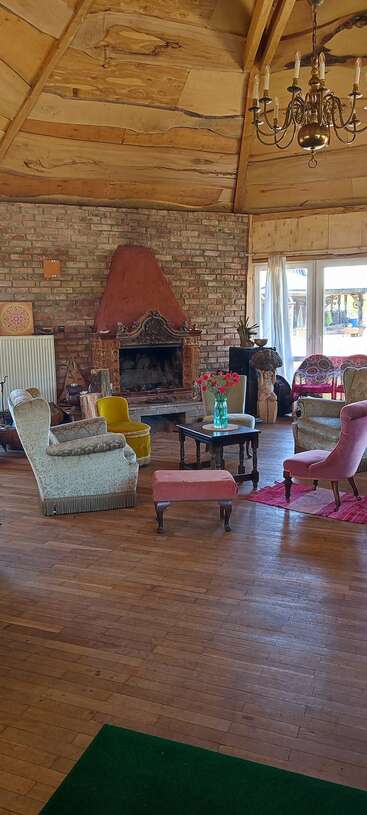 A cozy rustic living room features mismatched armchairs, a wooden ceiling, brick fireplace, chandelier, coffee table with flowers, and natural light from large windows.