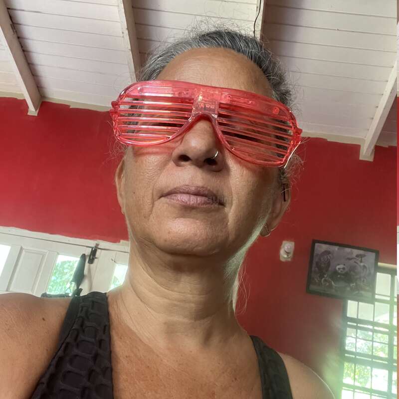 A woman with gray hair wears bright red shutter shades and a black top. She stands indoors against a red wall with a painting and white ceiling above.