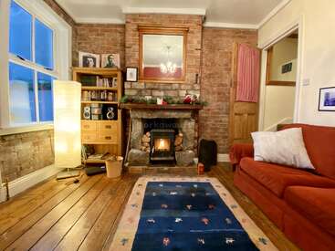 This cozy living room features a brick fireplace, wood-burning stove, red sofa, bookshelf, blue rug, standing lamp, wooden floors, and warm, inviting decor.