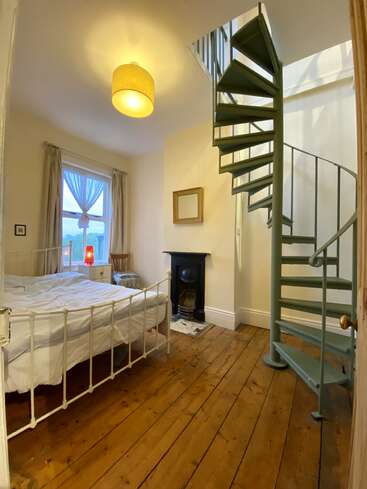 A cozy bedroom with a metal bed, wooden floor, fireplace, window with curtains, warm lighting, and a green spiral staircase leading to an upper floor.
