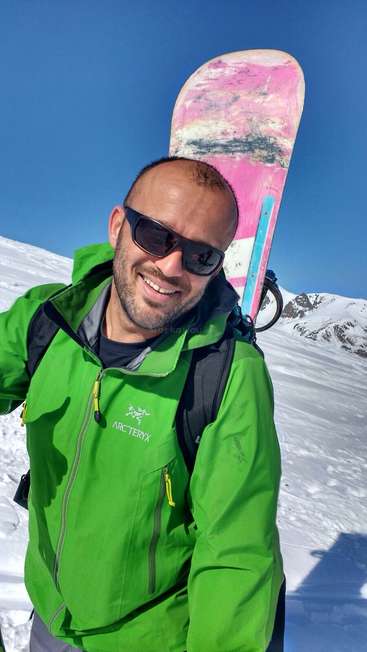 La imagen muestra a un hombre de pie en la nieve, con una chaqueta verde y una tabla de snowboard a la espalda, sobre un fondo montañoso nevado.