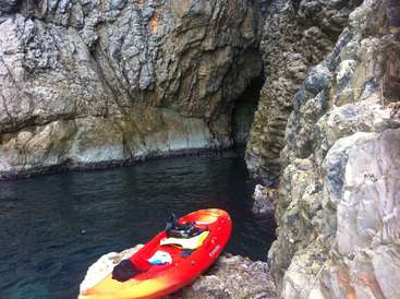 La imagen muestra un kayak rojo y amarillo situado sobre una roca cerca de una masa de agua, con un acantilado rocoso al fondo.