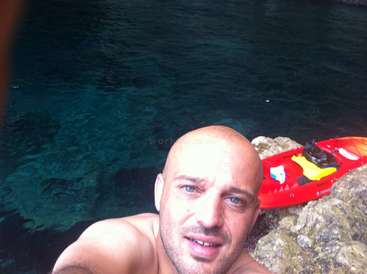 La imagen muestra a un hombre tomándose un selfie sobre una roca cerca del agua, con un kayak rojo detrás. El hombre está sin camiseta y tiene la cabeza calva.