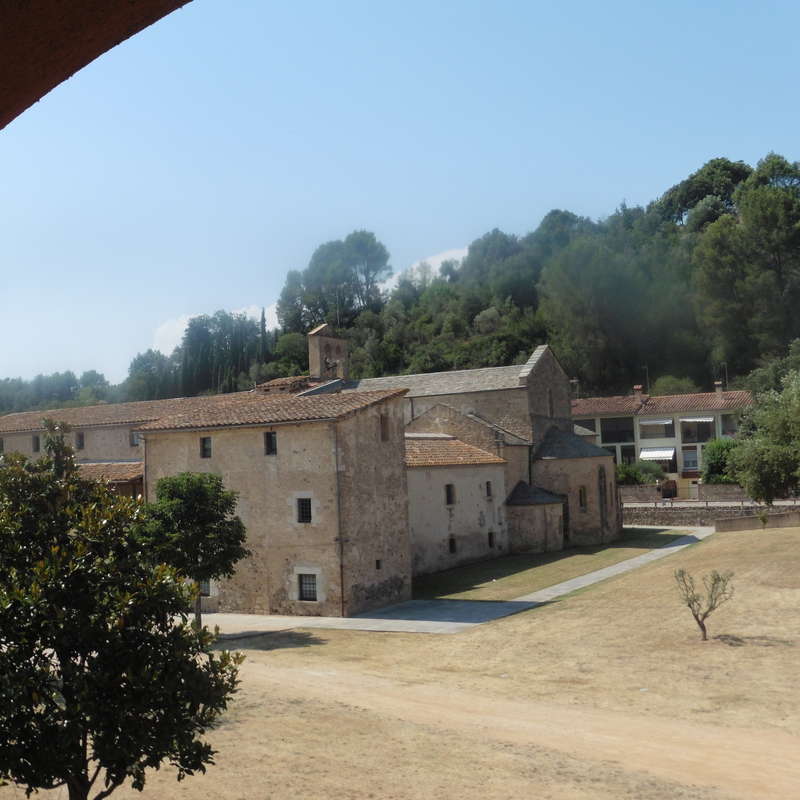 La imagen representa una serena escena rural con un gran edificio de piedra con tejado de tejas, rodeado de árboles y un cielo azul despejado en un día soleado.