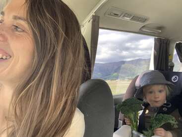 Una mujer sonriente conduce mientras un niño en un asiento de coche, con sombrero, sostiene brócoli fresco. A través de la ventanilla del vehículo se ven montañas y cielos nublados.