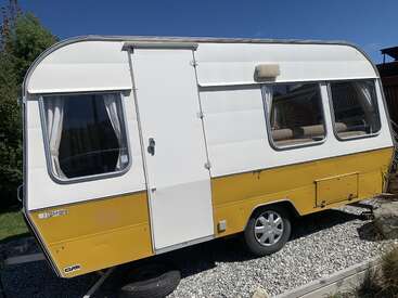 Una pequeña caravana vintage amarilla y blanca se asienta sobre la grava con la luz del sol reflejándose en sus ventanas. Detrás se ven árboles verdes y un cielo azul despejado.